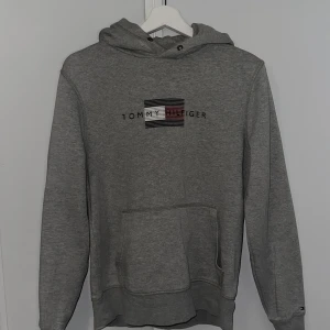 Grå hoodie från Tommy Hilfiger - Säljer en snygg och bekväm grå hoodie från Tommy Hilfiger. Den är i storlek S och gjord av en blandning av ekologisk bomull. Hoodien har en känguruficka perfekt för en avslappnad stil! Den är köpt i NK, nypris 1459 kr.