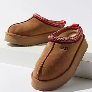 Bruna mockaskor från UGG - Säljer ett par supermysiga bruna mockaskor från UGG. De har en fluffig insida som håller fötterna varma och en snygg röd kant med mönster. Perfekta för kalla dagar när du vill vara både bekväm och stilren. Skorna har en robust sula som ger bra grepp.
