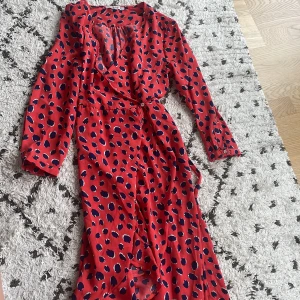 Röd omlottklänning med prickigt mönster - Säljer en superfin röd omlottklänning med blå prickar. Klänningen har långa ärmar och ett knytband i midjan. Perfekt för fest eller andra roliga tillfällen! 💃