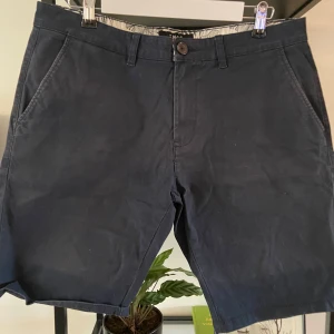 Svarta shorts från SMOG - Säljer ett par snygga mörk blåa shorts från SMOG i storlek L. De är perfekta för sommaren och har en klassisk design med två framfickor och två bakfickor. Materialet känns som bomull och de har en knapp och dragkedja framtill. Perfekta för både vardag och avslappnade tillfällen!