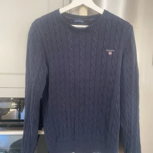 Mörkblå stickad tröja från GANT - Säljer en snygg och stilren mörkblå stickad tröja från GANT. Tröjan har ett klassiskt kabelstickad mönster och är gjord av bomull. Okej men använt skick. Perfekt för höst och vinter.