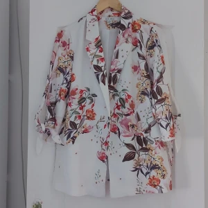 Blommig kavaj från Amisu - Säljer en superfin blommig kavaj från Amisu i storlek 38. Den är vit med ett färgglatt blommönster i rosa, röd, gul och brun.  Kavajen har 3/4 ärmar med slitsar som knyts. Har klassisk krage. Perfekt för vår och sommar! 🌸