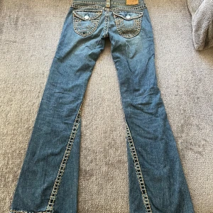  True Religion jeans - Säljer ett par snygga blå bootcut jeans från True Religion i storlek S. De har coola detaljer med kontrastsömmar och knappar på bakfickorna. Jeansen är i bra skick och perfekta för både vardag och fest. Passar dig som gillar en avslappnad men ändå stilren look. Köp direkt eller ge prisförslag! 💙Midjebredd är 35cm och innerbenlängd är 81 cm🩷