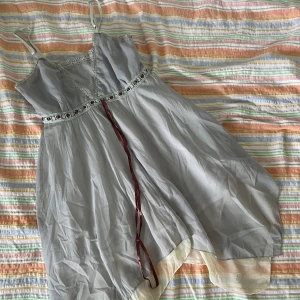 Ljusgrå klänning med spetsdetaljer - Säljer en superfin ljusblå asymmetrisk topp med spetsdetaljer och ett band i midjan. Klänningen har tunna axelband och är perfekt för sommarens alla fester. Den har en luftig passform.  