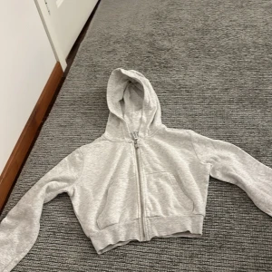 Grå croppad hoodie - Säljer en snygg och bekväm grå croppad hoodie. Den har en dragkedja framtill och en stor ficka på magen. Perfekt för en avslappnad stil eller för att hålla sig varm under kyliga dagar. Passar bra till både jeans och träningskläder. Köp direkt eller ge prisförslag 💕