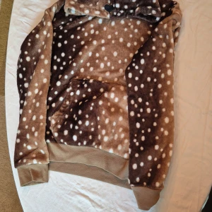 Brun fleece hoodie med hjortmönster från H&M - Säljer en supermysig brun fleece hoodie från H&M med ett gulligt hjortmönster. Den har en stor ficka på magen och en huva med öron och ögon, vilket gör den extra söt. Perfekt för kalla höst- och vinterdagar. Den är i bra skick och väldigt bekväm att ha på sig. 🦌