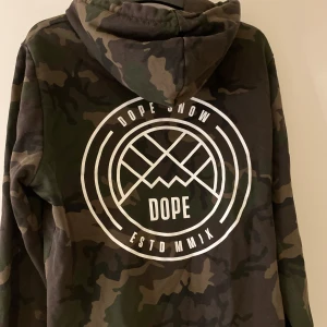 Camouflage hoodie från Dope - Säljer en snygg camouflage hoodie från Dope. Den har en stor vit logga på ryggen och en mindre på bröstet. Storlek är 40 men är som storlek S. Väldigt skön tröja nästan aldrig annvänd