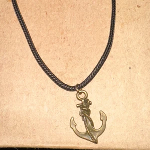 Halsband med ankare - Säljer ett coolt halsband med ett ankare som hänge. Kedjan är svart och flätad, vilket ger en snygg kontrast till det antikguldiga ankaret. Perfekt för en maritim look eller som en stilren accessoar till vardagsoutfiten.