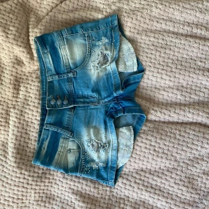 Blå jeansshorts med spetsdetaljer - Säljer ett par snygga blå jeansshorts från Denim Ax. De har coola slitningar och pärldetaljer på framsidan, samt en unik spetsdetalj på ena bakfickan. Shortsen har en låg midja och knappar framtill. Midjemått 34 cm, baksida 25 cm, framsida till gren 19 cm 💗