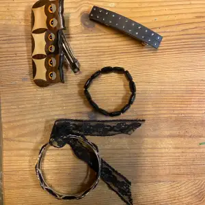 Ett coolt smyckeset som innehåller ett svart armband med ovala pärlor och ett silverfärgat armband med spetsdetaljer. Setet inkluderar också två hårklämmor, en i trä med ett unikt mönster och en svart med små glittriga stenar. Perfekt för att ge en extra touch till din outfit!