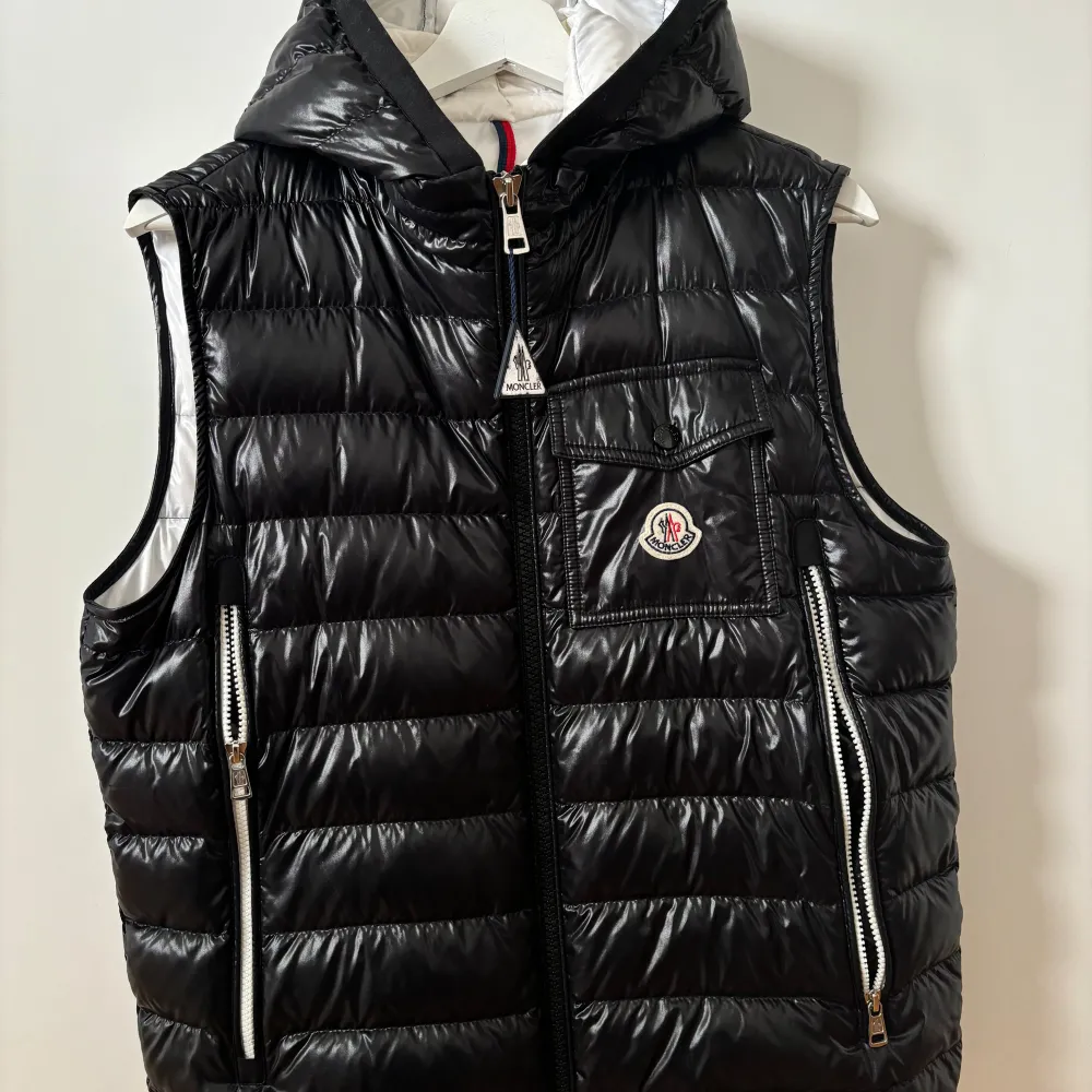 Säljer en snygg svart dunväst från Moncler. Västen har en quiltad design med dragkedja framtill och två praktiska fickor med dragkedjor. Den har också en liten bröstficka med Moncler-loggan. Insidan är vit och västen har en avtagbar huva. Perfekt för kyliga höst- och vinterdagar!. Jackor.