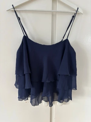 Marinblå volangtop från Gina Tricot - Säljer en marinblå topp från Gina Tricot med tunna axelband och flera lager volanger. Storlek 38🩷