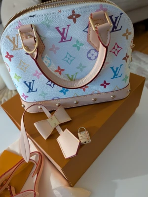 multicolor handväska - Handväska i vitt med färgglatt monogrammönster. Väskan har ljusrosa detaljer, guldfärgade metalldelar och medföljande axelrem, hänglås och nyckel. Perfekt för dig som vill sticka ut med en klassisk designerbag.