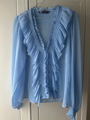 Ljusblå volangblus från Zara - Säljer en ljusblå blus från Zara med volanger längs den djupa v-ringningen och framtill. Blusen är gjord i ett tunt, transparent tyg och har långa ärmar med resår vid ärmsluten. Använd vid mycket få tillfällen💕💕