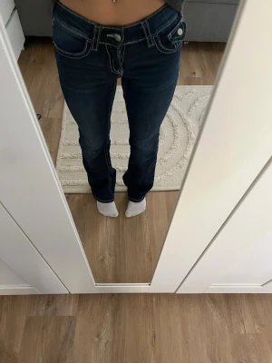Mörkblå bootcut jeans med vita sömmar - Snygga mörkblå jeans med tydliga vita kontrastsömmar och bootcut passform. Jeansen har låg midja och dekorativa fickor med lock bak. Perfekta för en trendig och avslappnad stil. Byxorna är i storlek 152.