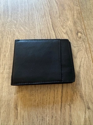 Svart plånbok i läderskinn - En stilren svart plånbok i skinn med klassisk vikbar design. Plånboken har flera fack för kort och sedlar, perfekt för att hålla ordning på dina viktigaste saker. Enkel och tidlös modell som passar till allt. I nyskick.