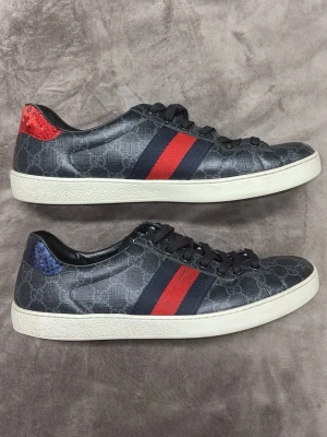 Gucci sneakers med röd och blå detalj - Snygga svarta Gucci sneakers med klassiskt GG-mönster, röda och blå ränder på sidan samt kontrasterande hälflikar i rött och blått ormskinn. Skorna har snörning och vit sula. Storlek 44-45