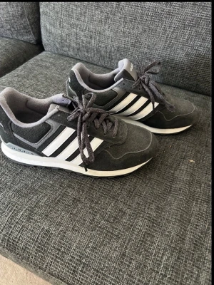 Gråa adidas sneakers med vita ränder - Gråa sneakers från adidas med klassiska vita ränder på sidorna. Ovandel i mocka och syntet, snörning och vit platt sula. Perfekt för dig som gillar sportig och stilren look.