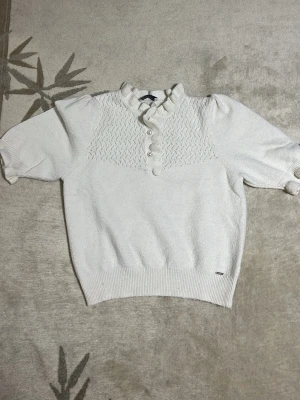Vit stickad topp med puffärm - Säljer en söt vit stickad topp med korta puffärmar och ribbade muddar. Toppen har ett dekorativt mönster upptill, liten ståkrage med volang och tre knappar framtill. Perfekt för dig som gillar en romantisk och trendig stil.
