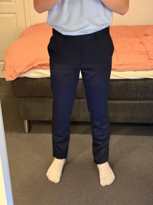 Mörkblå kostymbyxor med rak passform - Snygga mörkblå kostymbyxor med klassisk rak passform och bälteshällor. Byxorna har diskreta bakfickor med knappar och är perfekta för en clean och stilren look. Tillverkade i ett mjukt material som känns bekvämt hela dagen.