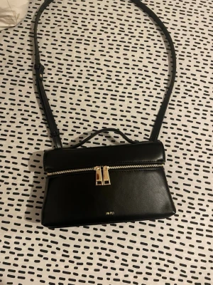 Svart handväska från JW PEI - Stilren svart handväska från JW PEI i slät syntet med justerbar axelrem och topphandtag. Väskan har guldfärgade detaljer, dragkedja och minimalistisk design. Perfekt för dig som gillar en clean och modern look.