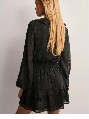 Svart volangklänning med lång ärm - Snygg svart kortklänning med volangdetaljer och v-ringning. Klänningen har långa ärmar, markerad midja med smock och dubbla volanger i kjolen. Tunt, lätt material med diskret mönster. Perfekt för dig som gillar trendiga och feminina plagg.