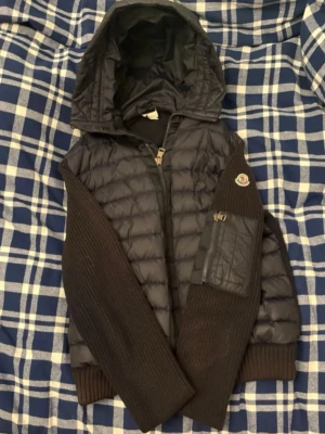 Svart cardigan jacka från Moncler - Svart cardigan jacka från Moncler med quiltad front och huva. Ärmarna är stickade och har en ficka med dragkedja på ena ärmen. Jackan har ribbade muddar och stängs med dragkedja framtill. Perfekt för lager-på-lager och snygg streetstil.