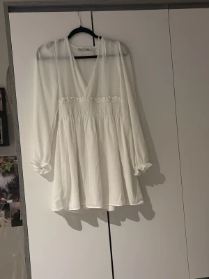 Vit omlottklänning med lång ärm - Säljer en söt vit kortklänning med omlott framtill och smock i midjan. Klänningen har långa, transparenta ärmar med resår vid ärmslut och ett luftigt, flowy tyg. Perfekt för sommarens alla tillfällen! 