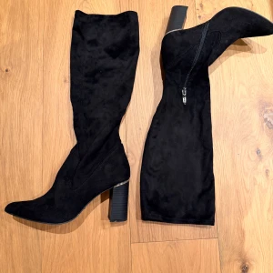 Svarta höga stövlar från Tamaris - Snygga svarta stövlar från Tamaris i mocka-look med hög klack och spetsig tå. Stövlarna har dragkedja på insidan och en stilren design som passar perfekt till höst- och vinteroutfits.