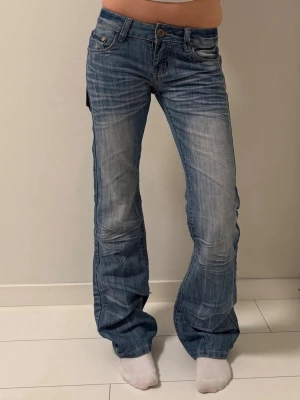 Blå bootcut jeans - Säljer ett par blå jeans med bootcut passform och snygga slitningar. Jeansen har låg midja, klassiska fem fickor och detaljerade sömmar på bakfickorna. 