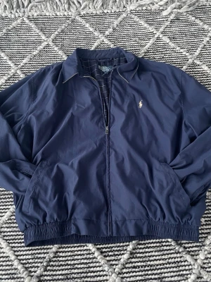 Marinblå jacka från Polo Ralph Lauren - Klassisk marinblå vindjacka från Polo Ralph Lauren med dragkedja framtill och broderad logga på bröstet. Jackan har krage, elastisk midja och muddar samt två sidofickor. Perfekt för vår och höst när du vill ha en stilren look. En innerficka finns. Mått kan skickas vid efterfrågan! Storlek XL men passar L också