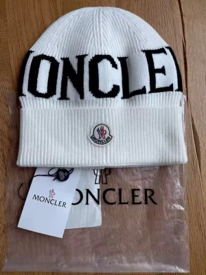 Vit Moncler mössa med logga - Säljer en stilren Moncler-mössa i vitt med stor svart logotext framtill. Klassisk ribbstickad modell som passar perfekt till både vardag och vinterfits. Mjuk och bekväm passform.  ✔️ Original enligt märkning ✔️ One size ✔️ Mycket fint skick ✔️ Kommer med tags och originalpåse (se bilder)