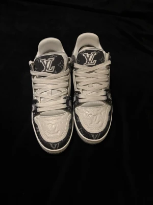 Louis Vuitton monogram sneakers med box och dustbags - Snygga Louis Vuitton sneakers i vitt och svart med klassiskt LV-monogram på ovandelen och präglad logga på tån. Skorna har vita snören, låg profil och robust sula med VUITTON-tryck undertill. Tillverkade i en mix av läder och textil för en exklusiv känsla. Storlek 45 men passar även 44