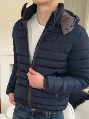 Mörkblå pufferjacka med huva - Snygg mörkblå pufferjacka med huva och dragkedja framtill. Jackan har två sidofickor med dragkedja och quiltad design. Perfekt för kyliga dagar och ger en sportig look. Fodrad insida och hög krage för extra värme.