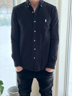 Svart skjorta från Ralph Lauren - Klassisk svart skjorta från Ralph Lauren med vit broderad logga på bröstet. Skjortan har långa ärmar, knappar framtill och button-down krage. Tillverkad i mjuk bomull som ger en skön känsla och passar perfekt till många olika stilar.