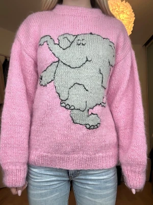 Rosa stickad tröja med elefantmotiv - Söt rosa stickad tröja med ett stort grått elefantmotiv på framsidan. Tröjan har rund halsringning, långa ärmar och är gjord i ett mjukt, fluffigt material som känns riktigt cozy. Perfekt för dig som gillar gulliga och lekfulla plagg.