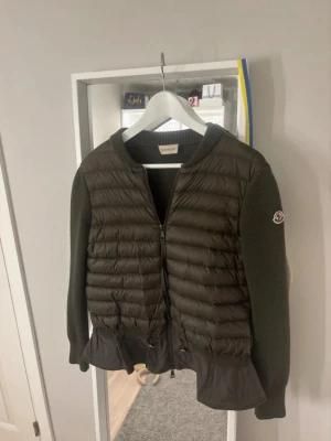 Grön cardigan jacka från Moncler - Snygg mörkgrön cardigan jacka från Moncler med quiltad framsida, stickade ärmar och rygg samt volangdetalj nedtill. Jackan har dragkedja framtill och Moncler-logga på ärmen. Perfekt för dig som vill ha en unik och trendig look.