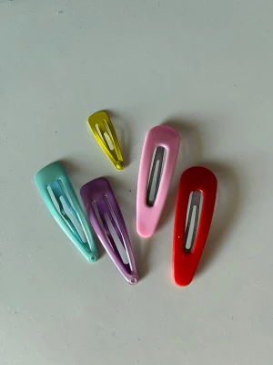 Färgglada klämmor i metall - Säljer ett set med fem hårspännen i olika färger: ljusblå, lila, rosa, röd och gul. Alla spännen har en klassisk klick-modell och är tillverkade i metall med blank finish. Perfekta för att piffa upp frisyren med en färgklick.