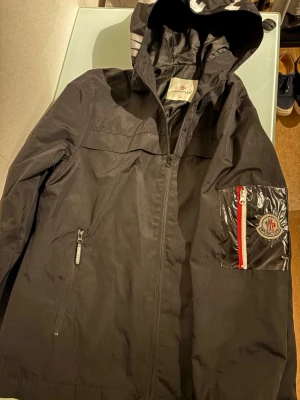 Svart vindjacka från Moncler - Säljer en svart vindjacka från Moncler med huva och dragkedja framtill. Jackan har en blank ficka på ärmen med rödvit dragkedja och Moncler-logga. Insidan har ett unikt serietryck. Perfekt för dig som vill ha en clean och sportig look.