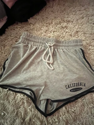 Grå shorts med snörning och tryck - Snygga grå shorts med svart kant och snörning i midjan. Tryck med texten 'California West Coast' på ena benet. Perfekta för sommaren och riktigt sköna att ha på sig. Tillverkade i mjuk bomullsmix.