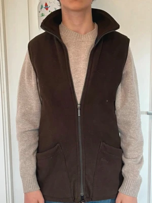 Mörkbrun fleeceväst från Barbour - Mysig mörkbrun fleeceväst från Barbour med två stora framfickor och dragkedja hela vägen. Insidan har klassiskt rutigt Barbour-foder och hög krage för extra värme. Perfekt att slänga över en tröja när det är kyligt ute.