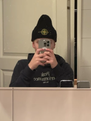 Svart ribbstickad mössa Stone Island - Svart ribbstickad mössa från Stone Island med klassisk logotyp-patch framtill i gult och grönt. Mössan har uppvikt kant och är tillverkad i ett mjukt, värmande material som passar perfekt för kyliga dagar.