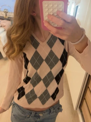 Beige argyle tröja med V-ringning, från Scandinavian golfer - Snygg beige tröja med klassiskt argylemönster i svart och grått framtill. Tröjan har V-ringning och långa ärmar, perfekt för en preppy look. Materialet ser ut att vara mjukt och stretchigt, vilket gör den bekväm att bära. 