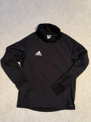 Svart Adidas träningströja med hög krage. - Svart långärmad träningströja från Adidas med vit logga på bröstet. Tröjan har hög krage och är tillverkad i ett lätt och funktionellt material, perfekt för sport och löpning. I mycket fint skick.