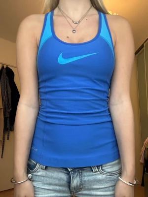 Blå träningstopp från Nike - Snygg blå träningstopp från Nike med ljusblå detaljer och klassisk logga på bröstet. Tight passform och brottarrygg. Perfekt för gymmet eller löprundan.