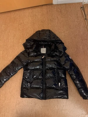 Svart pufferjacka från Moncler - Snygg svart pufferjacka från Moncler med glansig finish och stor huva. Jackan har två framfickor med dragkedja och quiltad design. Perfekt för kalla dagar när du vill hålla stilen. Klassisk Moncler-logga på ärmen och dragkedja framtill.