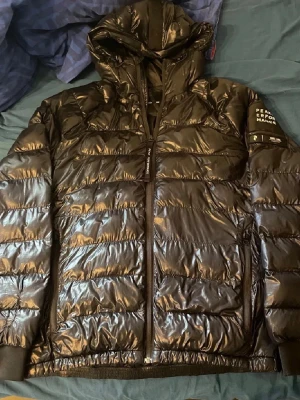 Svart dunjacka från Peak Performance - Snygg svart dunjacka från Peak Performance med glansig finish och huva. Jackan har dragkedja framtill, två sidofickor och logga på ärmen. Perfekt för kalla dagar och ger en modern look.