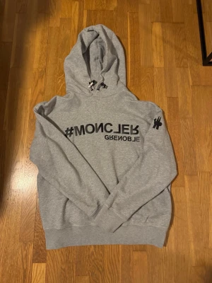 Grå Moncler hoodie med tryck - Snygg grå hoodie från Moncler Grenoble med svart tryck framtill och på ärmen. Klassisk huva med dragsko och ribbade muddar. Perfekt för en avslappnad och trendig stil. Mjukt material som känns skönt mot huden.