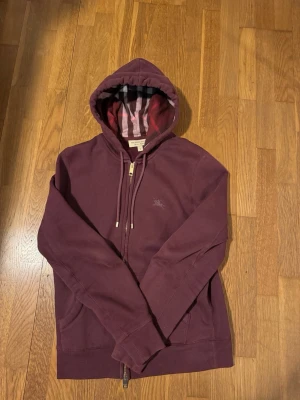 Vinröd hoodie från Burberry - Säljer en vinröd hoodie från Burberry med dragkedja och klassiskt rutigt foder i huvan. Hoodien har dragsko i huvan, två fickor fram och diskret Burberry-logga broderad på bröstet. Tillverkad i mjuk bomullsblandning för extra komfort. Öppen för att diskutera rimliga priser och tveka inte för att skriva.