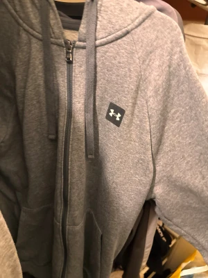 Grå zip hoodie från Under Armour / pris går att diskutera  - Snygg grå hoodie med dragkedja från Under Armour. Klassisk passform med huva och dragsnören, samt fickor framtill. Tillverkad i mjukt sweatshirtmaterial och har en diskret logga på bröstet. Perfekt för chill eller träning.